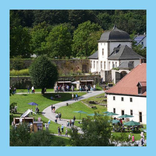Foto eingefasst von blauem Rahmen. Foto zeigt Blick auf das sommerliche Klostergelände mit Ständen und Besuchenden.