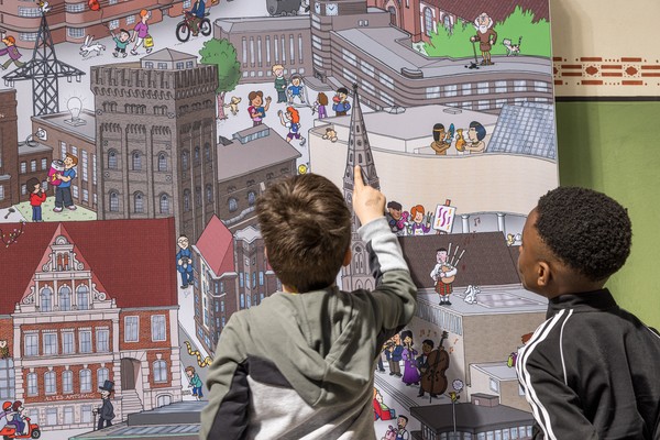 Zwei Jungen betrachten und zeigen auf ein großes Wimmelbild mit verschiedenen Gebäuden und vielen kleinen Figuren.
