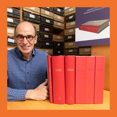 Mann in kariertem Hemd sitzt an Holztisch mit fünf dicken roten Büchern und Regal mit Archivboxen im Hintergrund. Foto mit einem orangenem Rahmen umrandet.