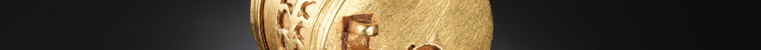 Ein zylindrisches Goldobjekt mit filigranen Gravuren und einem Schlüsselloch an einem Ende, auf einer reflektierenden schwarzen Oberfläche ausgestellt