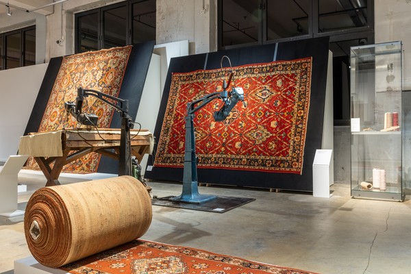 Industrielle Maschine mit Greifarm vor zwei an der Wand hängenden, gemusterten Teppichen und einer großen Teppichrolle auf dem Boden in einer Werkhalle