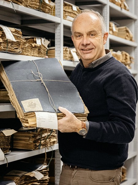 Mann in dunklem Pullover hält gebündelte alte Aktenordner in einem Archiv mit Regalen voller ähnlicher Aktenordner
