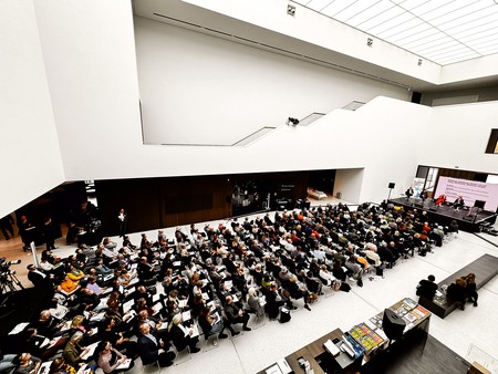 Großer Saal mit Publikum bei einer Konferenz vor einer Bühne mit drei sitzenden Personen und einer großen Leinwand