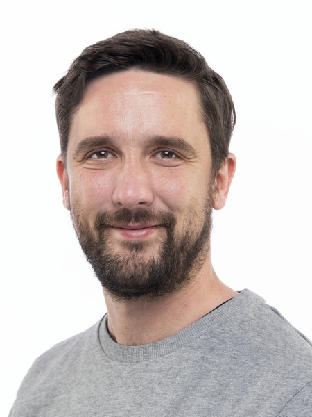 Mann mit kurzem dunklem Haar und Bart trägt graues T-Shirt und lächelt in die Kamera