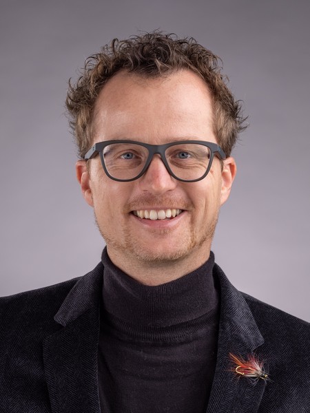 Lächelnder Mann mit lockigem Haar, Brille und schwarzem Rollkragenpullover unter schwarzem Sakko vor einfarbigem Hintergrund
