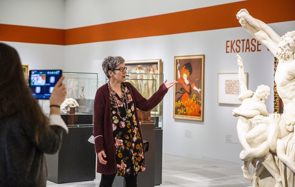 Person in floralem Kleid und rotem Cardigan zeigt auf weiße Skulptur in Museumsausstellung mit orangefarbenem Schriftzug 'EKSTASE' an der Wand