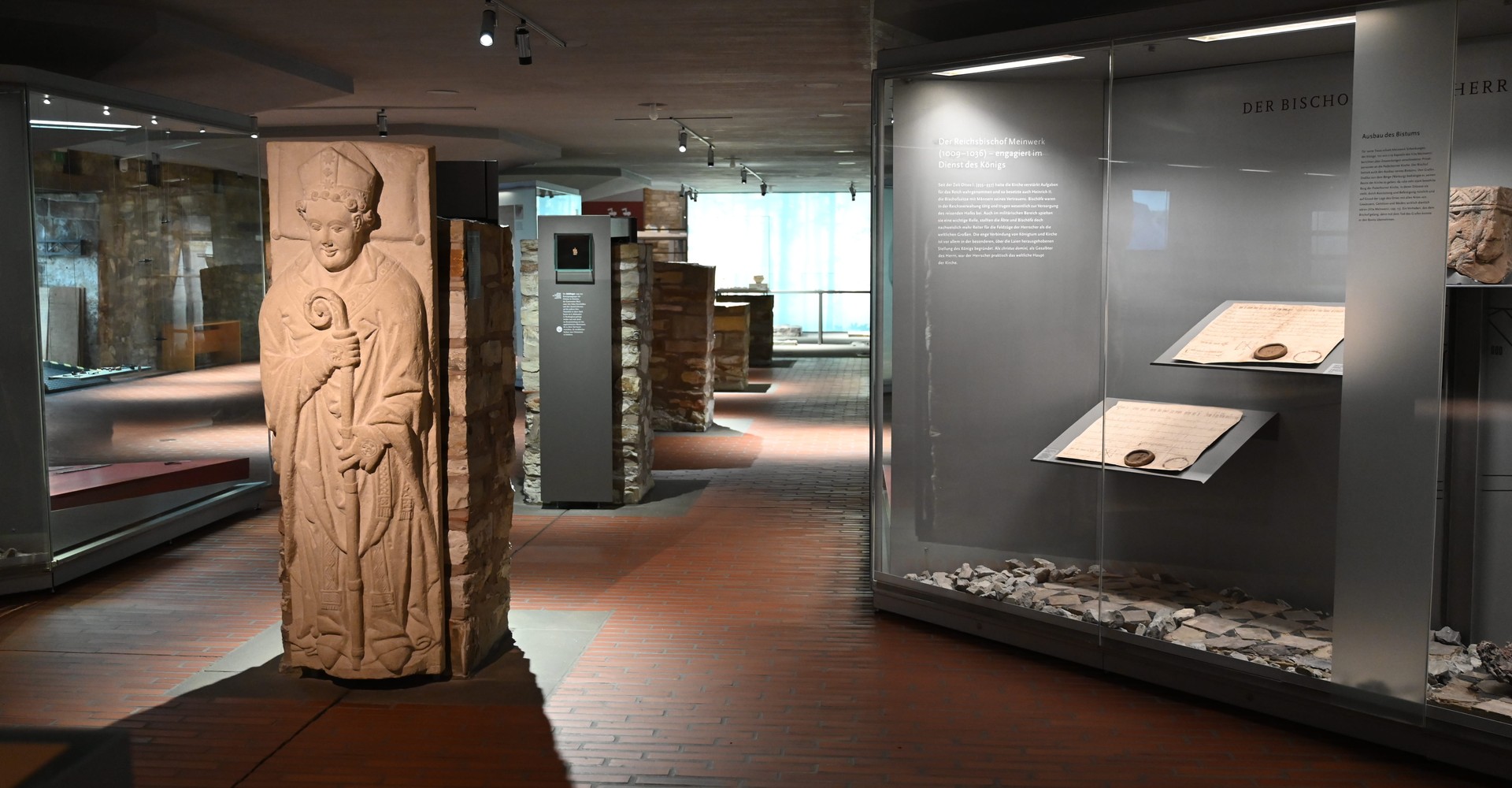 Museumsausstellung mit einer großen steinernen Skulptur einer stehenden Figur und Vitrinen mit historischen Artefakten.