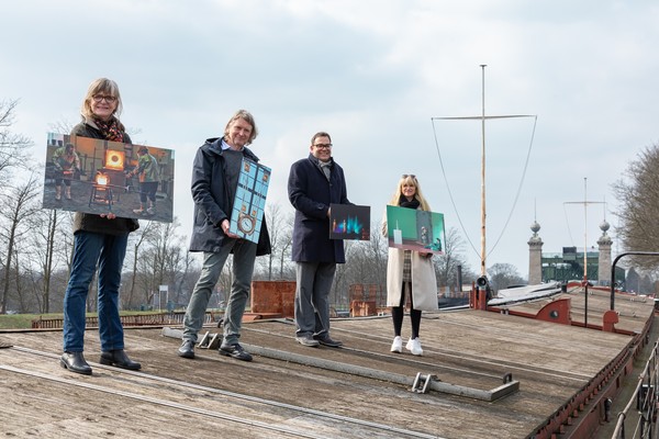Vier Personen stehen auf einem hölzernen Schiffsdeck und halten jeweils ein Bild mit unterschiedlichen Motiven in den Händen.