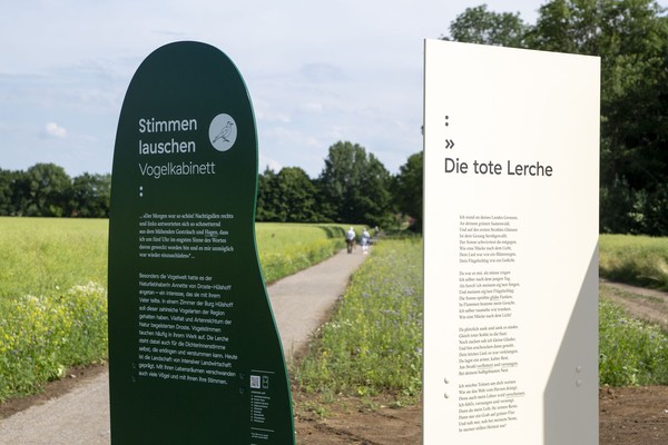 Zwei Informationstafeln am Wegesrand, eine dunkelgrün mit Text und Vogel-Illustration, die andere weiß mit Gedichttext, umgeben von Wiesen und Bäumen unter bewölktem Himmel.