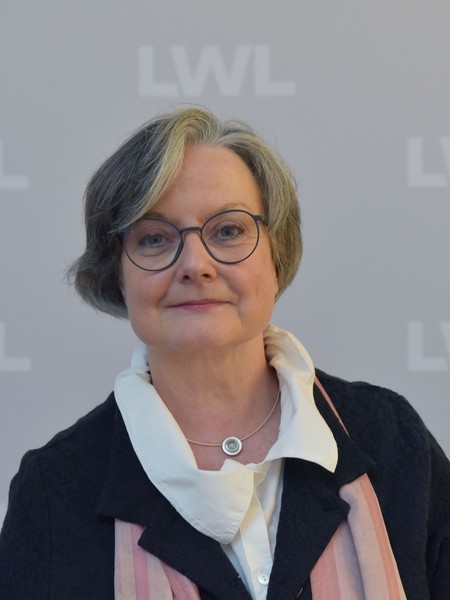 Frau mit Brille, grauem Haar und schwarzem Blazer vor grauem Hintergrund mit wiederholtem Schriftzug 'LWL'