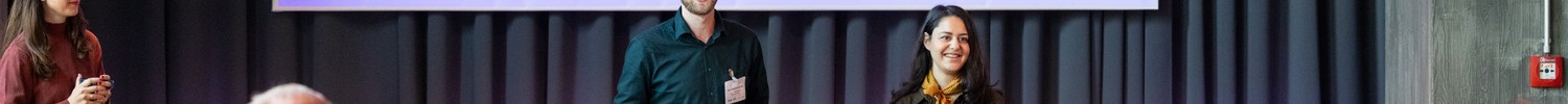 Drei Personen stehen vor einem Publikum und präsentieren vor einer Leinwand mit Programmablauf in einem Veranstaltungsraum