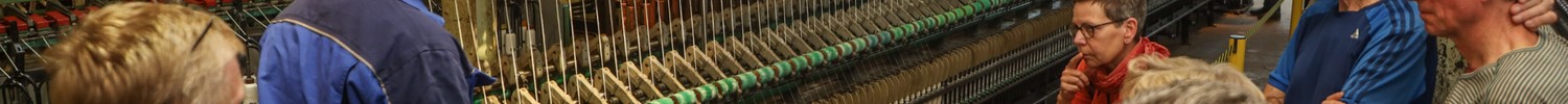 Gruppe von Menschen unterschiedlichen Alters steht vor einer langen Spinnmaschine in einer Fabrikhalle.