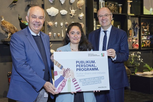Drei Personen in formeller Kleidung halten gemeinsam ein Plakat mit der Aufschrift 'PUBLIKUM. PERSONAL. PROGRAMM. Kultur divers und inklusiv LWL-MUSEUM FÜR NATURKUNDE' vor einer Wand mit naturkundlichen Exponaten wie Vogelschädeln und Muscheln