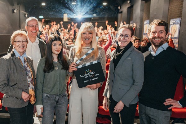 Fünf Personen stehen vorn in einem Kinosaal, eine hält eine Filmklappe mit der Aufschrift 'SCHULKINO', im Hintergrund sitzen viele Zuschauer auf roten Sitzen.