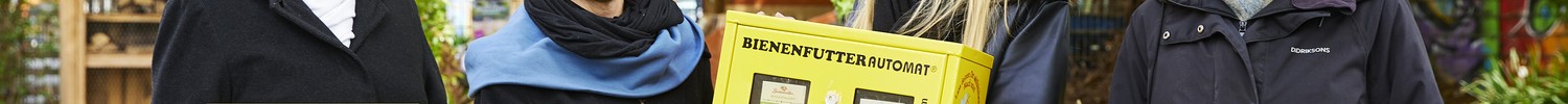 Vier Frauen stehen im Freien vor Bäumen und halten jeweils ein buntes Schild, eine gelbe Box mit der Aufschrift 'BIENENFUTTERAUTOMAT', ein kleines Vogelhaus und eine braune Tüte