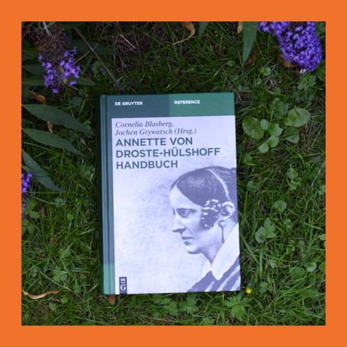 Buch mit dem Titel 'Annette von Droste-Hülshoff Handbuch' liegt auf Gras umgeben von lila Blüten. Foto ist von einem orangenem Rahmen eingerahmt.