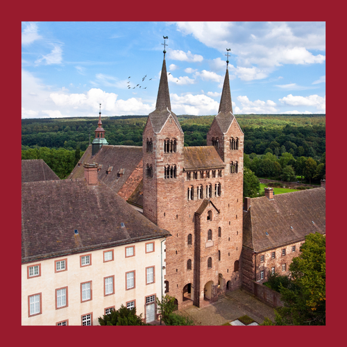 Historisches Klostergebäude mit zwei hohen Türmen und spitzen Dächern vor bewaldetem Hügeln. Foto eingerahmt von einem dunkelroten Rahmen.