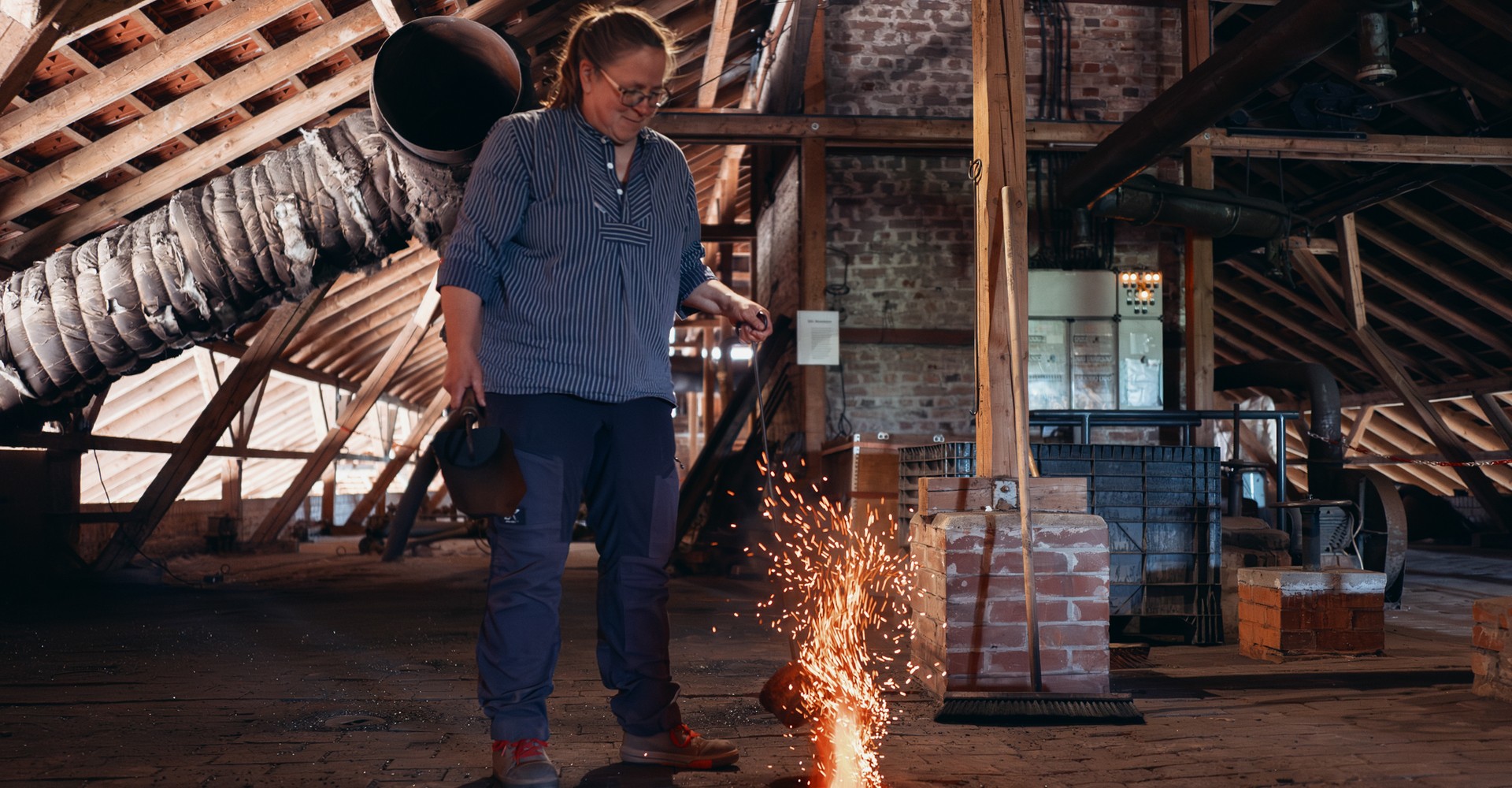 Person in Arbeitskleidung gießt geschmolzenes Metall in eine Form auf einem Boden aus Ziegeln in einer Werkstatt mit sichtbaren Holzbalken und Backsteinwänden.