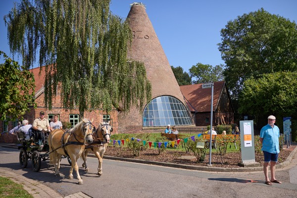 Zwei Pferde ziehen eine Kutsche vor einem Backsteingebäude mit kegelförmigem Dach und großem Bogenfenster bei sonnigem Wetter.