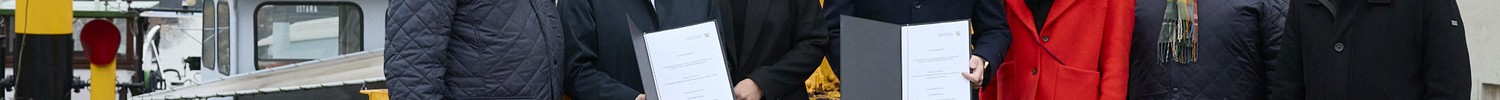 Sechs Personen stehen nebeneinander auf einem Kai vor einem gelben Hafenkran, zwei Personen halten jeweils ein Dokument in der Hand