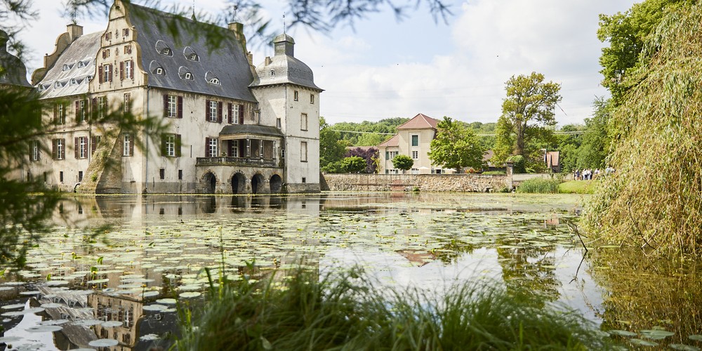 Wassergraben mit Seerosen vor einem historischen Schlossgebäude mit Giebeln und Türmen, umgeben von Bäumen und Wiesen