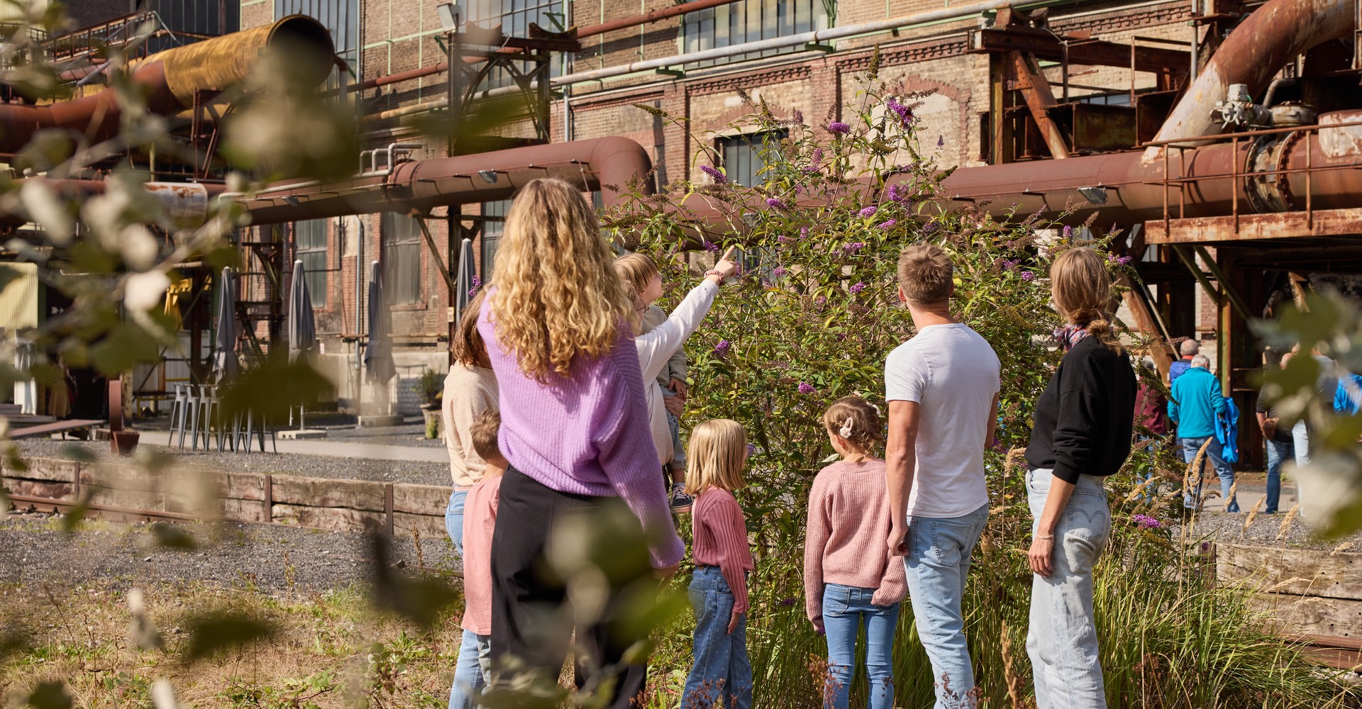 Gruppe von Kindern und eine erwachsene Person betrachten auf einer Industrieanlage ein Fliedergewächs.