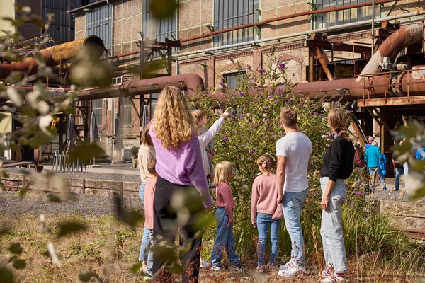 Gruppe von Kindern und eine erwachsene Person betrachten auf einer Industrieanlage ein Fliedergewächs.