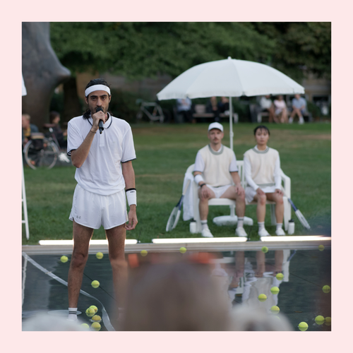 Ein Mann in weißer Tennisbekleidung steht auf einer Bühne im Freien und spricht in ein Mikrofon. Um ihn herum liegen zahlreiche Tennisbälle, während im Hintergrund zwei Personen unter einem Sonnenschirm sitzen und zuschauen.