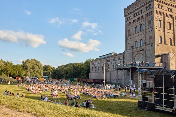 Menschen sitzen auf einer Wiese vor einem großen historischen Backsteingebäude unter blauem Himmel mit wenigen Wolken.