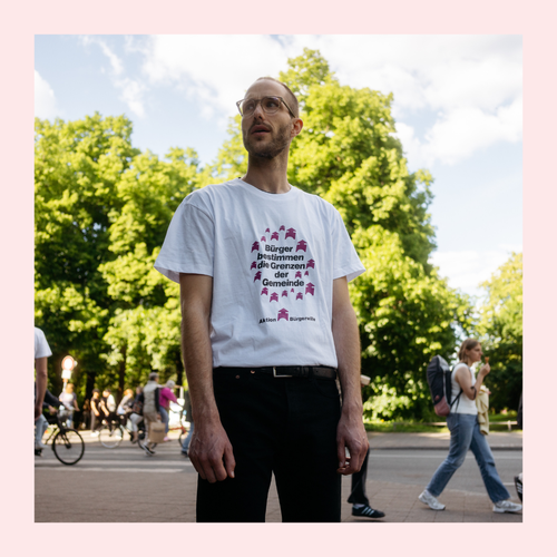 Mann mit weißem T-Shirt mit Aufdruck und schwarzer Hose steht auf einem Bürgersteig vor einer Straße, im Hintergrund mehrere Personen und grüne Bäume