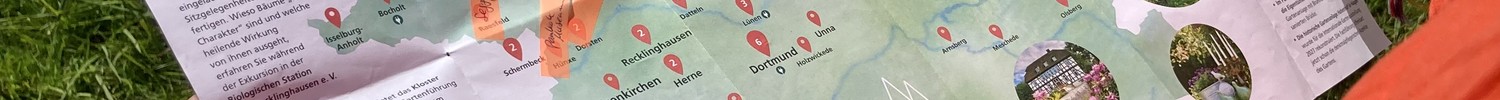 Person mit oranger Jacke und bunten Armbändern hält eine Karte mit einer Deutschlandkarte und markierten Orten auf grünem Gras