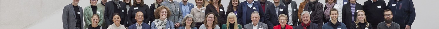Gruppenfoto von etwa 35–40 Erwachsenen in Business- und Smart-Casual-Kleidung mit Namensschildern, arrangiert in zwei Reihen auf einer Bank vor einer hellen, minimalistischen Betonwand mit Treppenstruktur, alle in die Kamera blickend.