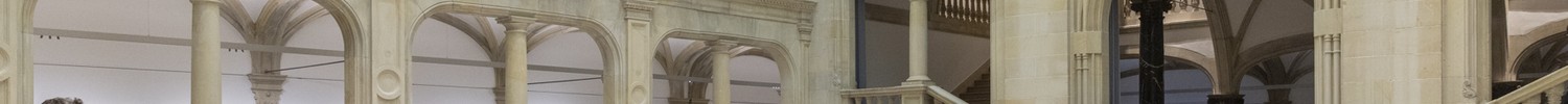 Menschen betrachten eine Ausstellung in einem großen historischen Innenraum mit Arkaden und einer Installation aus Holzbänken und Skulpturen.