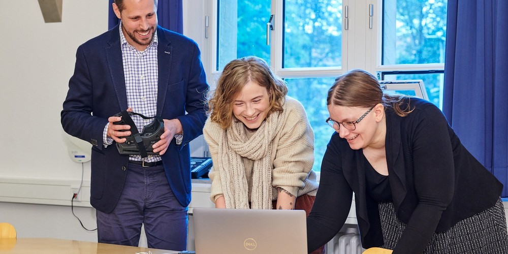 Drei Personen stehen an einem Tisch mit einem Laptop und elektronischen Bauteilen, eine Person hält ein VR-Headset