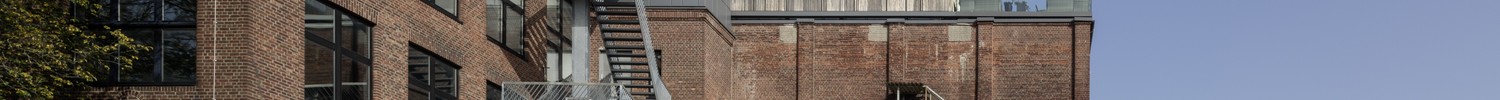 Backstein-Fabrikgebäude mit außenliegender Metall-Feuertreppe und teilweise abblätterndem Putz unter klarem Himmel, eine lange Rampe führt zur Eingang
