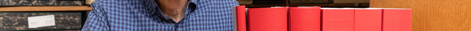 Person in blauem Hemd sitzt an einem Holztisch mit mehreren roten dicken Büchern vor sich und Archivregalen im Hintergrund