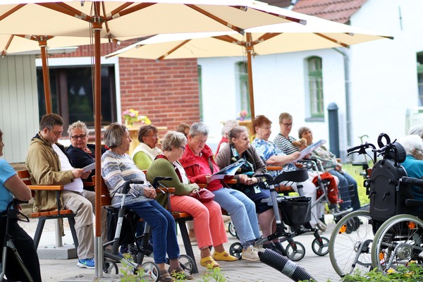 Gruppe älterer Menschen sitzt unter großen Sonnenschirmen auf Stühlen und Rollstühlen vor einem Gebäude