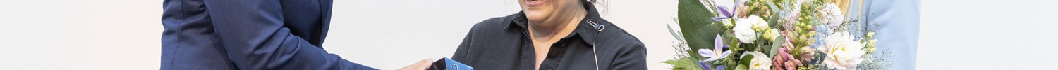 Eine Person überreicht auf einer Bühne eine Auszeichnung an eine Frau, während daneben eine weitere Person einen Blumenstrauß hält.