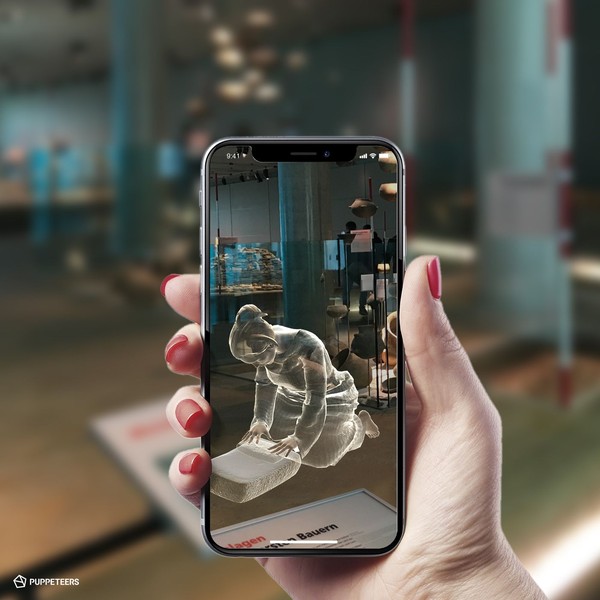 Hand hält ein Smartphone, das eine digitale Darstellung einer knienden Figur zeigt, im Hintergrund unscharfe Museumsausstellung
