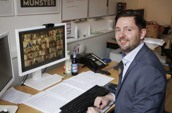 Person in Anzug sitzt an Schreibtisch mit Tastatur und Dokumenten vor einem Monitor, auf dem eine Videokonferenz mit vielen Teilnehmern zu sehen ist.