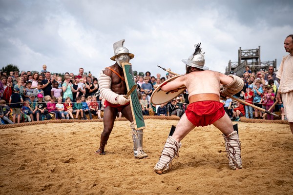 Zwei als Gladiatoren verkleidete Menschen stehen sich mit Waffe und Schild kampfbereit auf einem Sandfeld gegenüber, im Hintergrund eine Menschenmenge