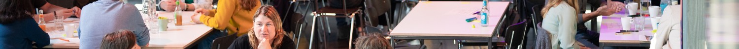 Mehrere Gruppen von Menschen sitzen an Tischen in einem Veranstaltungsraum mit Bühne und gelben Stühlen im Hintergrund