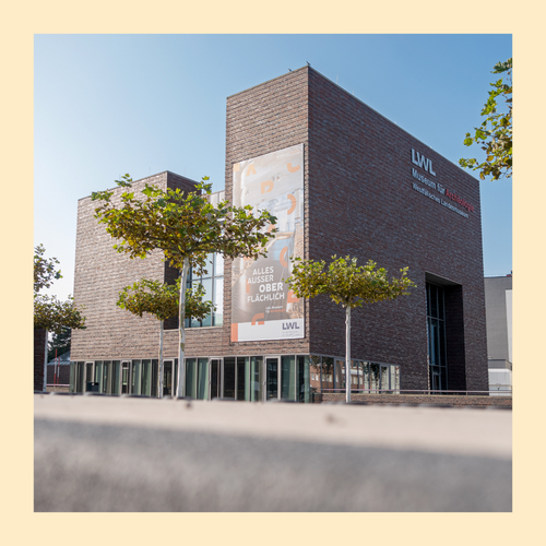 Modernes Backsteingebäude mit großen Fenstern und Banner mit Text 'ALLES AUSSER OBER FLÄCHLICH' vor blauem Himmel und Bäumen. Foto eingefasst in gelben Rahmen.