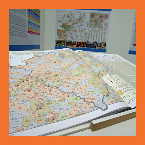 Aufgeschlagene Landkarte mit farblich markierten Regionen und Symbolen auf einem Tisch vor einer Wand mit weiteren Karten und Texten. Foto umrandet mit orangenem Rahmen.