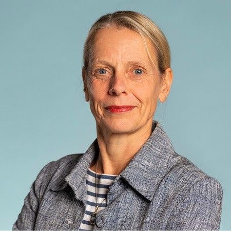 Frau mit blondem Haar im Dutt trägt graue Jacke und gestreiftes Shirt vor einfarbigem hellblauem Hintergrund