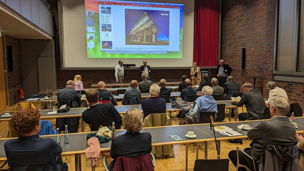 Mehrere Personen sitzen in einem Seminarraum mit Tischen und hören vier Vortragenden vor einer Leinwand mit einer Architekturpräsentation zu.