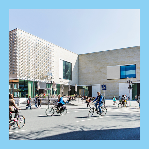 Eckiges helles Gebäude mit Installation an der Fassade, davor eine mit Fahrrädern befahrene Straße. Foto eingefasst in einen blauen Rahmen.
