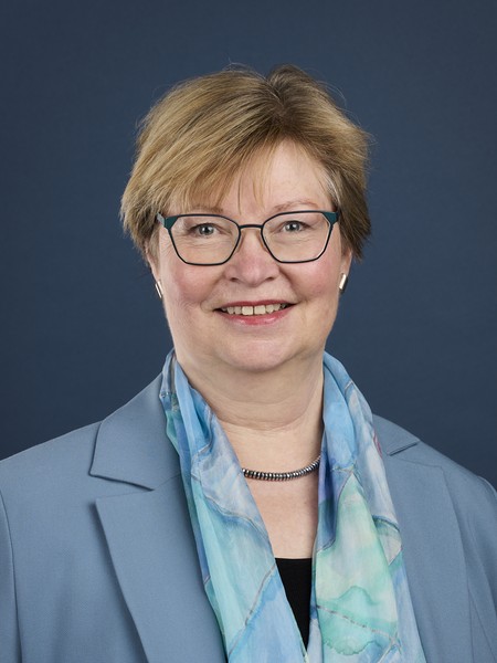 Frau mit Brille, kurzem blondem Haar, blauem Blazer und blau-grünem Schal vor dunkelblauem Hintergrund