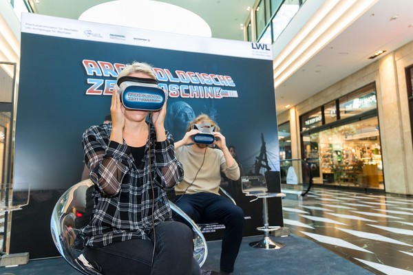 Zwei Personen sitzen auf transparenten Stühlen und tragen Virtual-Reality-Brillen vor einem Werbeplakat mit der Aufschrift 'ARCHÄOLOGISCHE ZEITMASCHINE'