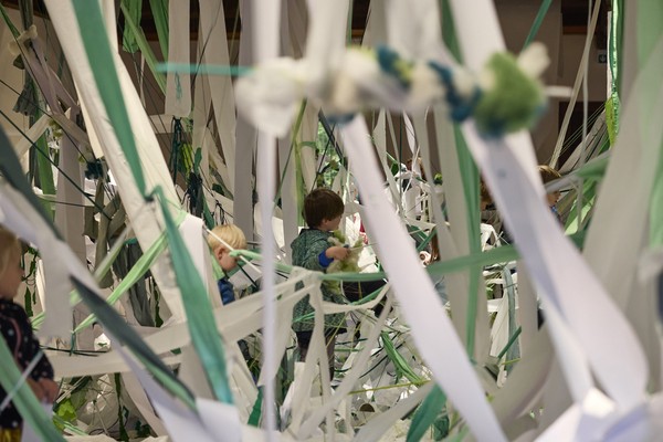 Zwei Kinder spielen zwischen vielen hängenden weißen und grünen Papierstreifen.